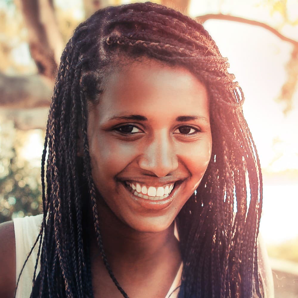 Portrait standing in for Trisha Brown: a smiling young woman with braids, in sunlight.